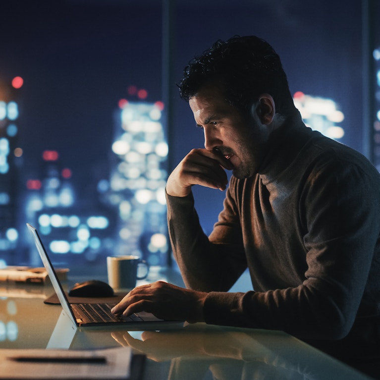Portrait of thoughtful successful businessman working on laptop computer in his big city office at night.