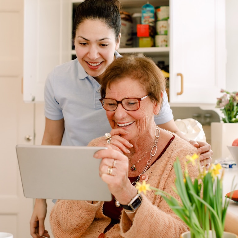 Smiling elderly woman and young female caregiver looking at digital tablet in nursing home kitchen