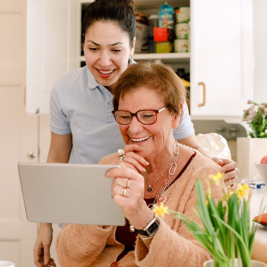 Smiling elderly woman and young female caregiver looking at digital tablet in nursing home kitchen
