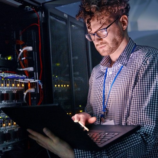 Focused male IT technician working at laptop in dark server room